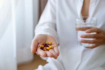 Closeup image of a sick woman holding pills and a glass of water while sitting on a bed