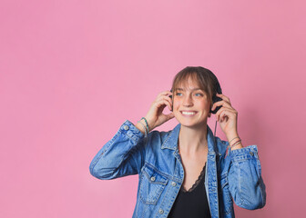 Studio portrait of a woman putting on headphones, she is listening to music and is smiling, happy and full of emotions. Standing in the center of the image, in front of a pink background.