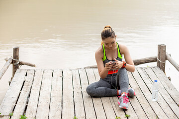 Young Asian woman in exercise clothes, wearing headphones, listening to music on smartphone, sitting by the waterfront.