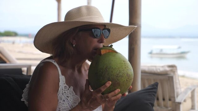 Caucasian middle-aged woman enjoying the life by drinking coconut milk from the coconut on the beach watching the ocean