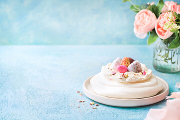 Meringue nest cake Pavlova with colorful chocolate eggs, buttercream frosting, light blue background with copy space. Selective focus. Food ideas for kids. Easter Spring love feast concept. Close up.