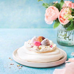 Meringue nest cake Pavlova with colorful chocolate eggs, buttercream frosting, light blue background with copy space. Selective focus. Food ideas for kids. Easter Spring love feast concept. Close up.