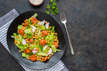 A healthy salad of green lettuce, radishes, tomato with boiled chickpeas on a black plate on a dark concrete background. Served with natural yoghurt sauce. Salad recipes.