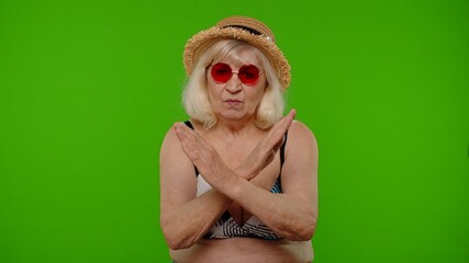 Dislike gesture. Bad idea. Wrong choice. Refusal signal. Senior woman traveler in red sunglasses showing stop sign. Mature grandmother in swimsuit on chroma key background. Tourism, summer vacation