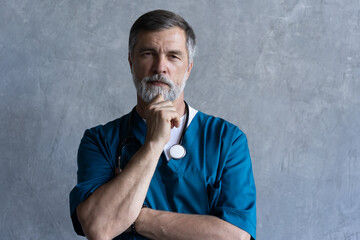 Portrait of professional mature surgeon looking at camera while standing against the grey background.