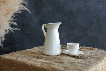 White decanter, white cup on white saucer, stand on burlap bedding on blue textured background and dry reed branches