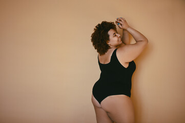 Fototapeta na wymiar Beautiful curvy oversize African black woman afro hair posing in black bodysuit on beige brown background isolated, body imperfection, body acceptance, body positive and diversity concept. Copyspace.