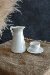 White decanter, white cup on white saucer, stand on burlap bedding on blue textured background and dry reed branches