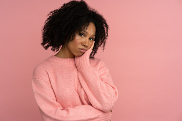 Melancholic sad African woman looking at camera with thoughtful expression. Depressed Afro-American girl keep hand on cheek, has skeptical expression, wear pink sweater isolated on pink studio wall. 