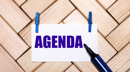 On a wooden background, a white card with the text AGENDA on blue clothespins and a blue marker. View from above