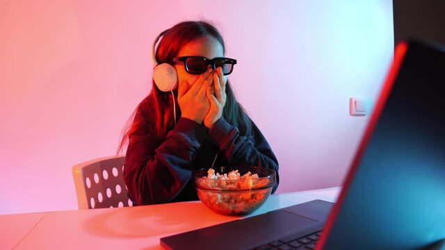 girl in glasses 3D and headphones watches terrible movie on laptop, eats popcorn