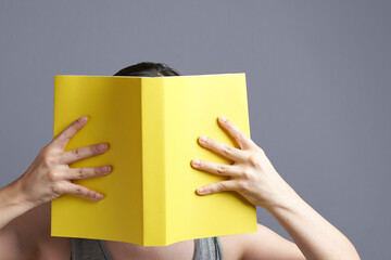 Young woman with her face hidden behind an open yellow book that she is reading