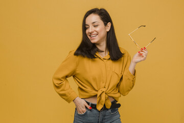 Photo of young female holds sunglasses and looks so happy. Wears yellow shirt, isolated yellow color background