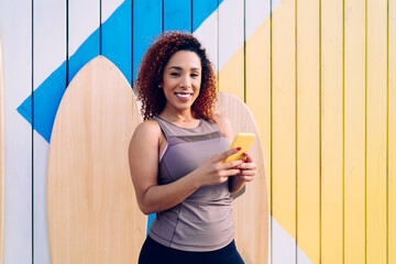 Cheerful sportswoman surfing smartphone while standing against colorful plank wall