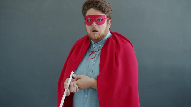 Portrait of funny actor in red superman costume licking huge lollipop and screaming looking at camera on gray background. Emotions and expressions concept.