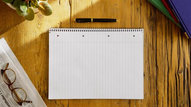 Top view shot of a blank page of a notebook with a person putting down cup of coffee on a dark, rough, wooden table. Newspaper, books, fountain pen and sunlight on background. 4k Close-up