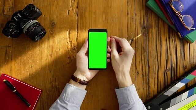 Top View of Filmmaker using Smartphone with Green Screen. Sunlight on Dark Coarse Wooden Desk. Vintage SLR Camera, Slate, Fountain Pen, Books on background. Touching, Zooming Gestures. 4k Close-up