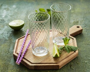 Two empty glasses, mint sprigs and lime slices on a wooden board against a green concrete background.