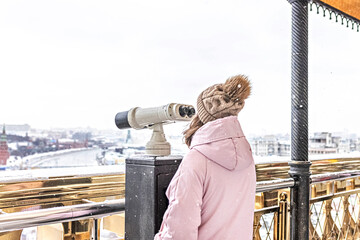 A young girl looks through coin-operated binoculars on the observation deck overlooking the city from a height at sunset. Winter, snowfall