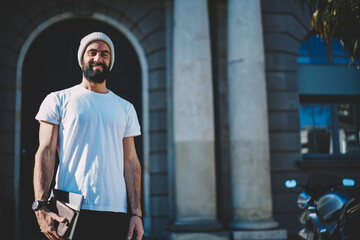 Half length portrait of cheerful Turkish student in hat smiling at camera holding education equipment in hand, happy Middle Eastern male with textbooks posing at street urban setting on leisure