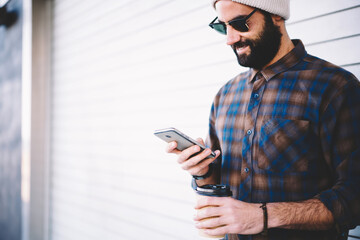 Middle Eastern hipster guy in sunglasses using mobile technology for making online booking and baking during travel vacations, millennial blogger with beard and coffee to go messaging via cellular