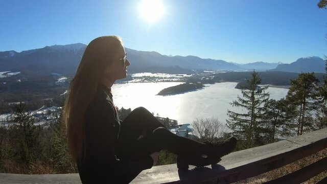 A woman in black sitting on a wooden rail and flipping her long hair with the panoramic view on Faak lake in Austrian Alps. The picturesque lake is surrounded by high mountains. Sun is shining bright.