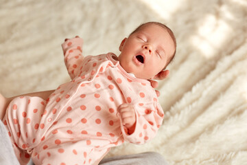 Sleeping newborn baby in mother hands, cute kid wearing sleeper with polka dots, kid keeps mouth opened, little girl in mommy's arms in background of white fluffy blanket.