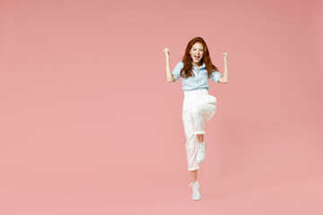 Fototapeta na wymiar Full length young student successful overjoyed happy redhead woman in blue shirt pants do winner gesture clench fist celebrate with raised up leg isolated on pastel pink background studio portrait