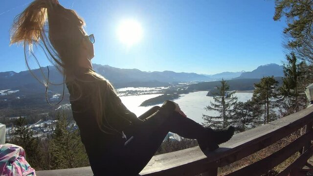 A woman in black sitting on a wooden rail and flipping her long hair with the panoramic view on Faak lake in Austrian Alps. The picturesque lake is surrounded by high mountains. Sun is shining bright.