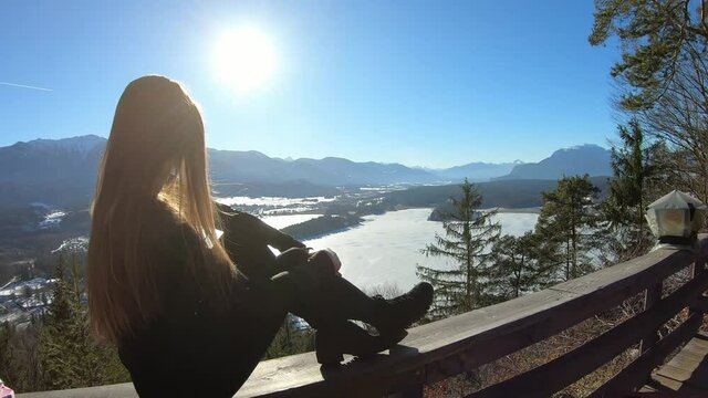 A woman in black sitting on a wooden rail and flipping her long hair with the panoramic view on Faak lake in Austrian Alps. The picturesque lake is surrounded by high mountains. Sun is shining bright.