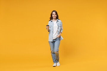 Full length of young smiling friendly fun student happy woman 20s in casual denim shirt white t-shirt holding paper cup of coffee drinking hot tea isolated on yellow color background studio portrait