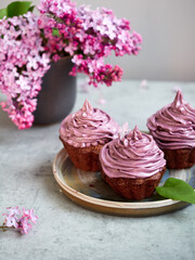 Baking, lilac, spring. Delicious chocolate cupcakes with lilac cream on top. Filled with chocolate. Surrounded by fresh lilacs. On a concrete countertop. Gray background. Space for the text. Content.