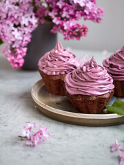 Baking, lilac, spring. Delicious chocolate cupcakes with lilac cream on top. Filled with chocolate. Surrounded by fresh lilacs. On a concrete countertop. Gray background. Space for the text. Content.