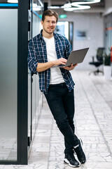 Caucasion male freelancer is standing in modern office, looking at the camera, smiling. Happy young adult businessman in stylish wear is using laptop, working on new project