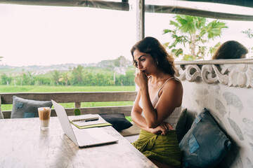 Pensive ethnic distance worker watching laptop on cafeteria terrace