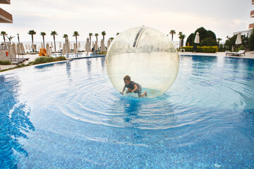 Little boy have fun inside big plastic balloon on the water of swimming pool on the summer resort. Kid inside big inflatable transparent ball running and having fun.