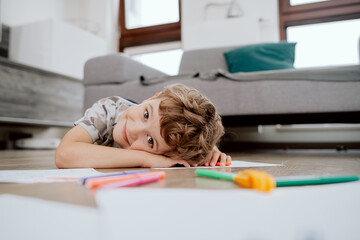 Portrait of school-age boy lies on the living room floor and paints pictures on white sheets of paper with colored markers. The boy is spending his afternoon free time at home.