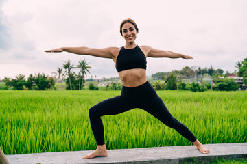 Fototapeta premium Portrait of cheerful woman in tracksuit doing Warrior Pose during yoga exercises in morning, female gymnastic enjoying virabhadrasana during time for physical recreation near Indonesian rice fields