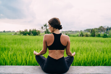 Fototapeta na wymiar Back view of active female dressed in black sportswear sitting in lotus pose near Indonesian rice fields and getting inspiration from nature, healthy woman recreating during hatha yoga training
