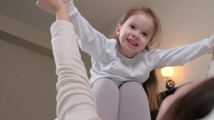 A kid, a girl, plays, laughs and flies on plane in her mothers arms. A child and a young mother play an airplane together on the couch at home, dreams of flying. Happy family. Family doing gymnastics