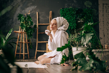 Beautiful woman in white bathrobe and towel sitting next to the bath in interior decorated with plenty of green plants - obrazy, fototapety, plakaty