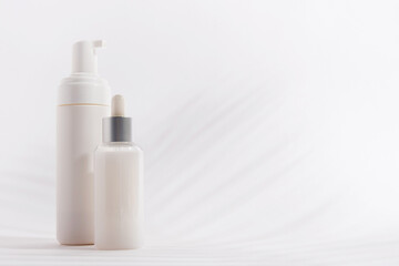 A container of mousse and a bottle of serum on a light background with shadows from a palm tree.