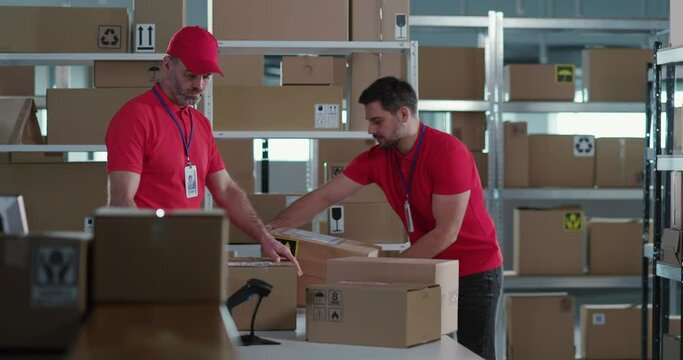 Caucasian bearded couple of manager and postal deliveryman wearing red uniform collecting and sorting delivery parcel boxes inside storage room. Post office.