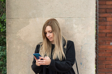 Mujer joven rubia con ojos claros mirando el teléfono móvil en una pared marrón 