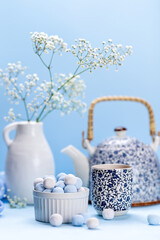 Teapot in a blue flower, cups, white vase with flowers and round sweets candies on a blue background. Blue still life with utensils.
