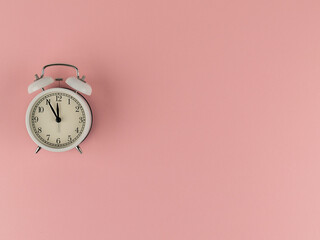 White retro alarm clock shows about twelve o'clock on pink paper background with copy space. Time:...