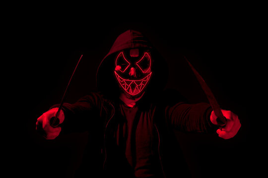 Man armed with two machetes and wearing a scary lighting neon glow mask and a hoodie on black background.