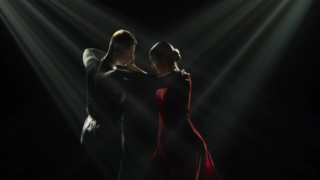 Dark silhouettes of professional ballroom dancers dancing Argentine tango in a dark studio with bright rays of light. Passionate glances, touching the hands and bodies of a man and a woman. Close up.