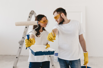 Happy young married couple in love in white T-shirts in yellow protective rubber gloves and glasses doing repairs updating painting the walls roller preparing to move into a new home, selective focus