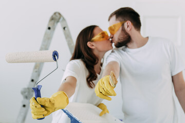 Happy young married couple in love in white T-shirts in yellow protective rubber gloves and glasses doing repairs updating painting the walls roller preparing to move into a new home, selective focus